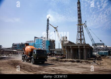 Betonbau, gelbe Mobilkrane, orange Betonmischer, blaues Industriegebäude, Fabrikschornsteine am blauen Himmel. Renovierungsarbeiten Stockfoto