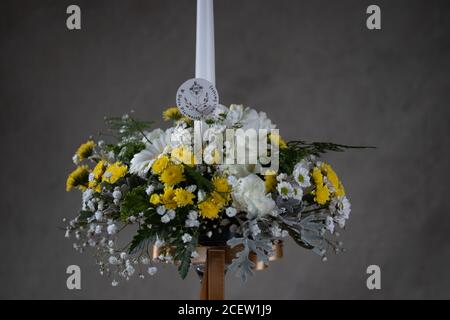 Erste heilige Kommunion Blumensträuße. Weiße Kerze mit weißen und gelben Blumen verziert. Graues Backgroud Stockfoto