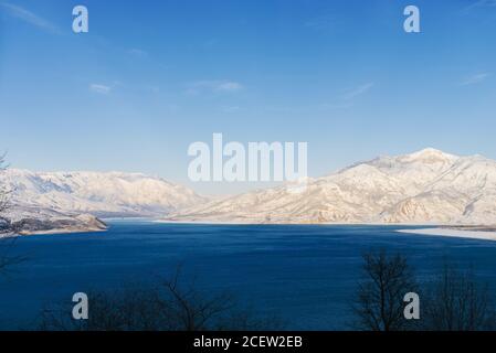 Charvak-Stausee mit blauem Wasser an einem klaren Wintertag in Usbekistan, umgeben von schneebedeckten Berggipfeln des Tien Shan Stockfoto