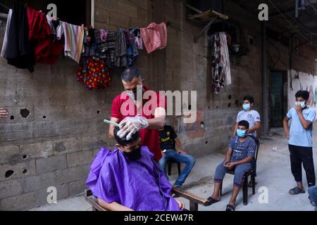 31. August 2020: Gaza, Palästina. 31. August 2020. Die Kinder halten etwas Abstand, während sie auf ihre Reihe warten, als ihnen der palästinensische Barbier Sameh Al-Qassas in der engen Gasse vor seinem Haus in Gaza City einen Haarschnitt gibt. Sameh trägt Masken und Handschuhe und desinfiziert ständig seine Werkzeuge und verteilt Handdesinfektionsmittel. Der drastische Anstieg der Covid-19-Infektionen seit der Entdeckung der ersten Gemeinschaftsfälle Anfang letzter Woche in Gaza hat die palästinensischen Behörden alarmiert, die eine strikte Sperre verhängt haben und die Bewohner aufgefordert haben, sich im dicht besiedelten Streifen sozial zu distanzieren. Stockfoto