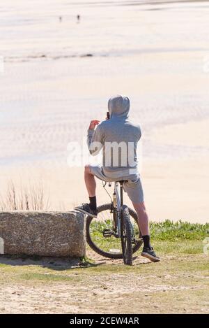 Ein Mann mit Hoodie sitzt auf seinem Mountainbike mit Blick auf Fistral Beach in Newquay in Cornwall. Stockfoto