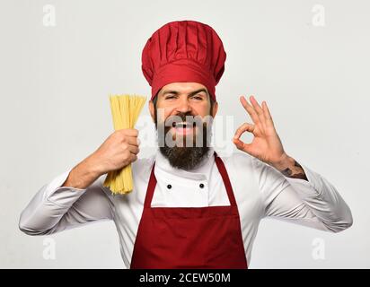 Cook mit zufriedenen Gesicht in burgunderroten Uniform hält trockene Pasta zeigt ok Zeichen. Der Koch hält einen Haufen Spaghetti in der Hand. Konzept der italienischen Küche. Mann mit Bart isoliert auf weißem Hintergrund. Stockfoto