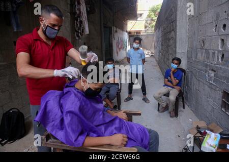 31. August 2020: Gaza, Palästina. 31. August 2020. Die Kinder halten etwas Abstand, während sie auf ihre Reihe warten, als ihnen der palästinensische Barbier Sameh Al-Qassas in der engen Gasse vor seinem Haus in Gaza City einen Haarschnitt gibt. Sameh trägt Masken und Handschuhe und desinfiziert ständig seine Werkzeuge und verteilt Handdesinfektionsmittel. Der drastische Anstieg der Covid-19-Infektionen seit der Entdeckung der ersten Gemeinschaftsfälle Anfang letzter Woche in Gaza hat die palästinensischen Behörden alarmiert, die eine strikte Sperre verhängt haben und die Bewohner aufgefordert haben, sich im dicht besiedelten Streifen sozial zu distanzieren. Stockfoto