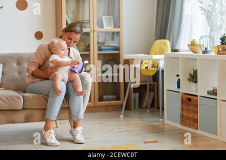 Horizontale lange Aufnahme der modernen kaukasischen Frau tragen lässige Outfit sitzen auf dem Sofa mit ihrem Kind auf dem Schoß spielen mit Spielzeug, kopieren Raum Stockfoto