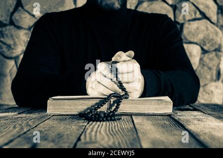 Mann in schwarzer Kleidung mit einem Rosenkranz in den Händen vor einem offenen alten Buch bibel oder koran, das Konzept des Gebets, monochromes Foto Stockfoto