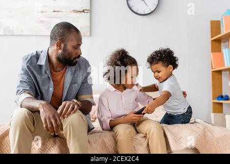 afroamerikanischer Mann in legerer Kleidung auf dem Bett in der Nähe sitzen Kinder mit fernbedienung Stockfoto