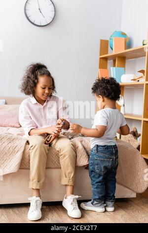 afroamerikanische Mädchen in legere Kleidung auf dem Bett sitzend und Spielzeug in der Nähe des kleinen Bruders halten Stockfoto