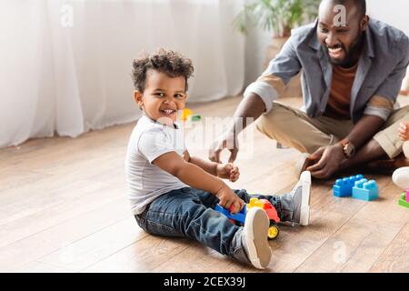 Aufgeregt afroamerikanischen Mann in der Nähe Sohn spielt mit Spielzeug LKW Auf Holzboden Stockfoto