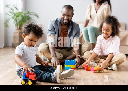 Selektiver Fokus auf afroamerikanischen Mann und Kinder spielen mit Die Gebäude auf dem Boden in der Nähe der Mutter, die auf dem Bett sitzt Stockfoto
