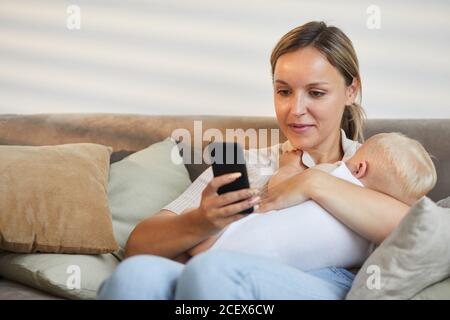 Moderne kaukasische Frau sitzt auf dem Sofa mit ihrem Smartphone während Kuscheln ihres kleinen Sohnes in den Armen Stockfoto