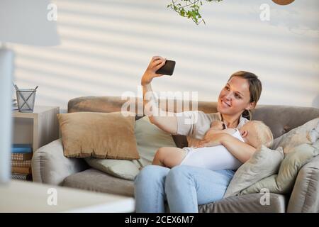 Moderne kaukasische Frau sitzt auf dem Sofa und nimmt Selfie-Foto auf Smartphone beim Kuscheln ihres kleinen Sohnes in den Armen Stockfoto