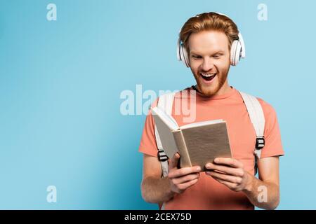 Begeisterter Schüler in drahtlosen Kopfhörern Buch auf blau lesen Stockfoto