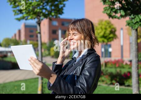 Glückliche junge stilvolle Frau im Park mit Tablet und Telefonieren Stockfoto