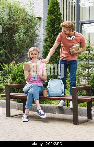 Rotschopf Student berühren blonde Freundin Buch lesen, während sitzend auf Bank Stockfoto