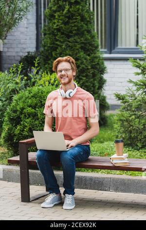 Bärtiger Student sitzt auf der Bank mit Laptop in der Nähe von Büchern und Papierbecher Stockfoto