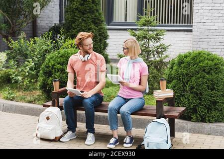 Bärtige und blonde Studenten sitzen auf einer Bank in der Nähe von Büchern, Pappbechern und Rucksäcken, während sie digitale Tablets halten Stockfoto