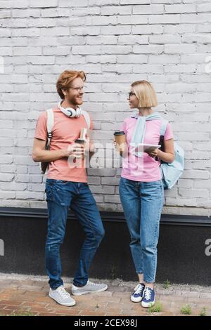 Studenten in Gläsern halten Kaffee zu gehen und zu sehen Einander, während sie in der Nähe der Ziegelmauer stehen Stockfoto