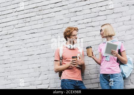 Junge Studenten in Gläsern halten Kaffee zu gehen und suchen Einander aneinanderstehend in der Nähe der Ziegelmauer Stockfoto