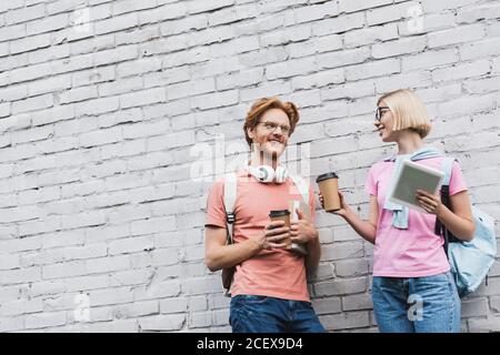 Junge Studenten halten Kaffee, um zu gehen und schauen auf jeden Andere, während in der Nähe der Ziegelmauer stehen Stockfoto