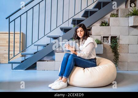 Volle Länge der fokussierten jungen weiblichen Designerin in lässiger Kleidung Sitzen auf bequemen Hocker und mit Grafik-Tablet während der Arbeit In modernen kreativen Arbeitsbereichen Stockfoto