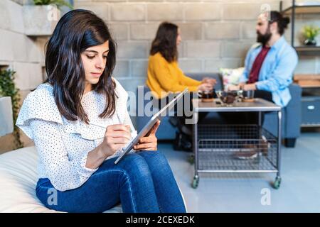 Serious junge weibliche Designerin in casual Outfit arbeiten an Projekt Mit modernem Grafiktablett, während Sie im kreativen Arbeitsbereich mit sitzen Kollegen im Hintergrund Stockfoto