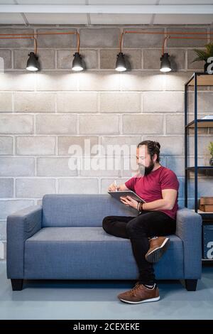 Voller Körper fokussierten jungen bärtigen männlichen Freiberufler in casual Outfit sitzen auf dem Sofa und mit Tablet während der Arbeit an Projekt im zeitgenössischen Loft-Arbeitsbereich Stockfoto