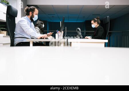 Ernst junger Mann in medizinische Maske mit Handy, während Kolleginnen, die mit Computern im modernen Coworking Office arbeiten Stockfoto