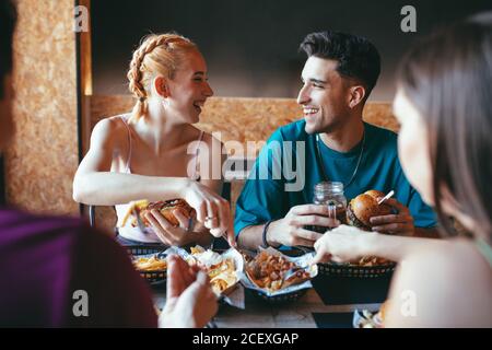 Fröhlicher junger Mann und Frau reden und lachen, während sie sich mit Freunden in der Cafeteria treffen und Fastfood-Snacks und Getränke genießen Stockfoto