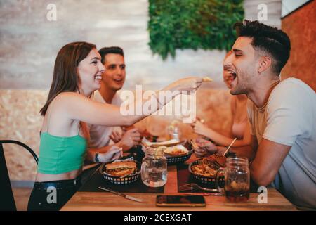 Fröhliche junge Frau, die einem lachenden Freund einen Snack gibt, während sie mit Freunden in der Cafeteria zu Mittag gegessen hat Stockfoto