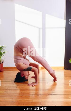 Seitenansicht einer folligen Frau, die in Salamba Sirsasana Yoga praktiziert Auf Holzboden zu Hause an sonnigen Tagen Stockfoto