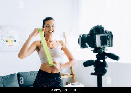 Selektiver Fokus der Sportlerin, die das Widerstandsband in der Nähe der Digitalkamera hält Auf Stativ zu Hause Stockfoto