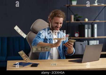 Gewinne online. Gewinnen Sie das Geld. Ich bin reich. Porträt eines jungen Freiberuflers oder Geschäftsmannes in einem Heimbüro. Wirft Bargeld. Erfolgreicher Abschluss Stockfoto