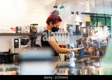 Seitenansicht der weiblichen Barkeeper in Gesichtsmaske stehen Theke im Coffee Shop und machen leckere heiße Getränke in Kaffeemaschine während der Coronavirus-Epidemie Stockfoto