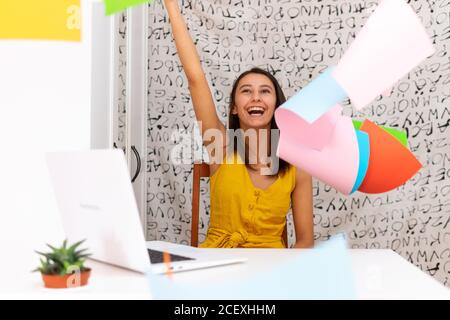 Entzückte Freiberuflerin sitzt am Tisch mit Laptop zu Hause Beim Feiern des Sieges und Werfen von bunten Papieren in der Luft Stockfoto