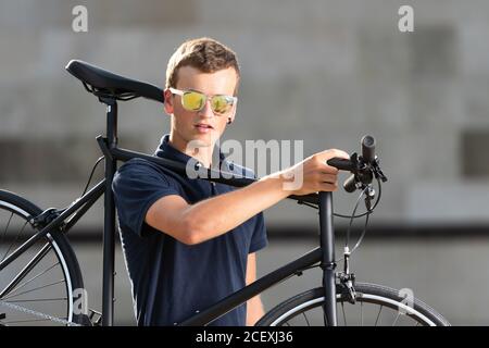 Junger Mann mit Sonnenbrille, der sein Stadtfahrrad auf der Schulter trägt Auf der Straße mit Blick auf die Kamera Stockfoto