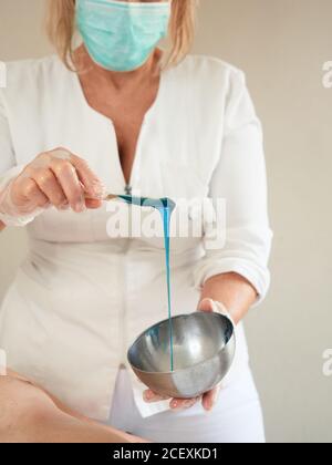 Gesichtslose Frau in medizinischer Maske und Handschuhe, die heißes blaues Wachs mit Holzstab in der Schüssel rühren Stockfoto