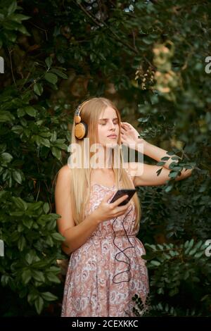 Heitere junge blonde Frau im Sommerkleid Musik hören Mit Smartphone und Kopfhörer zwischen grünen Laub im Garten mit Augen geschlossen Stockfoto