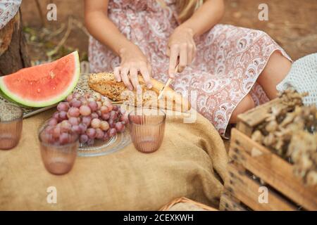Crop anonyme Frau im Sommerkleid schneiden knusprigen Brot Laib Auf dem Tisch mit frischem Obst und Glas Tassen während des Picknicks In der Natur Stockfoto