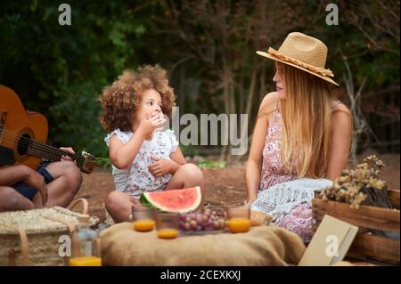 Liebenswert lockig behaart kleines Mädchen essen frische Früchte während genießen Sommerpicknick mit multirassischen Eltern im grünen Wald Stockfoto