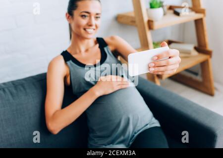 Selektiver Fokus der schwangeren Sportlerin beim Berühren Bauch während der Selfie Mit Smartphone auf Couch Stockfoto
