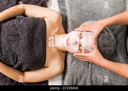 Von oben Ernte anonyme Kosmetikerin Anwendung Gesichtstuch Maske auf Weibliche Klientin während der Verjüngung Hautpflege Verfahren im modernen Schönheitssalon Stockfoto