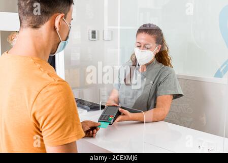 Junge Fachärztin in Schutzmaske hält Kartenleser während Männlicher Patient für die Behandlung mit Kreditkarte in modernen bezahlen Klinik Stockfoto