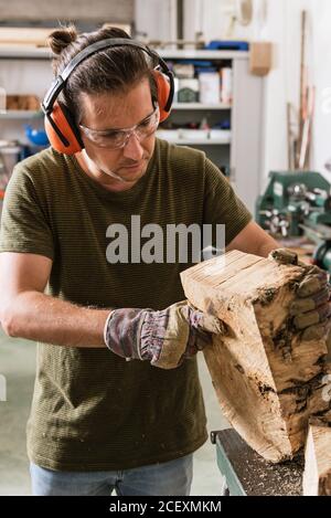 Konzentrierter männlicher Holzarbeiter in Schutzbrillen und industriellen Ohrenschützern Stehen an der Werkbank mit Holz Stockfoto