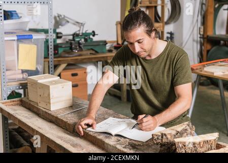 Konzentrierter männlicher Tischler, der an der Werkbank steht und sich Notizen machen kann Notebook während der Arbeit in der Garage Stockfoto