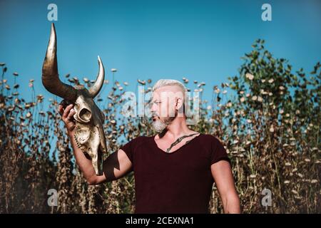 Serious muskulösen bärtigen grauhaarigen Mann mit Tattoo halten Horn Tierschädel beim Stehen gegen hohes Gras und blauen Himmel In der Natur Stockfoto