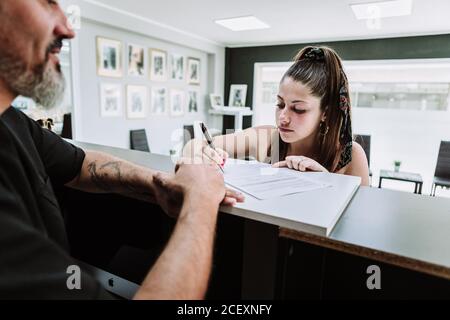 Junge Frau, die im Tattoo-Salon am Tresen steht und unterschreibt Vor dem Verfahren dokumentieren Stockfoto