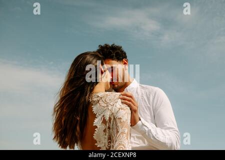 Von der unteren Seitenansicht der fröhlichen jungen Braut und des Bräutigams In stilvollen Hochzeitskostümen stehen umarmt einander mit Augen Geschlossen auf blau klaren Himmel genießen einander Stockfoto