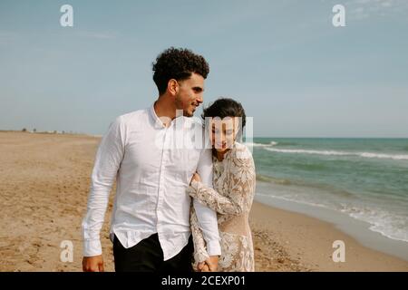 Glückliche junge Braut und Bräutigam in eleganten Boho Outfits halten Hände und umarmen einander mit geschlossenen Augen beim Spaziergang Entlang Sandstrand mit wehenden Meer Stockfoto