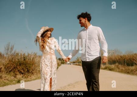 Glücklicher junger Mann in stilvollem Outfit, der die Hand der Frau im weißen Hochzeitskleid hält, während er mit fliegendem Flugzeug auf der Landstraße gegen den blauen Himmel schlendert Stockfoto