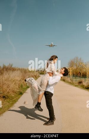 In voller Länge Seitenansicht von glücklichen jungen Mann in stilvollem Outfit trägt fröhliche Frau in weißem Hochzeitskleid, während auf Landstraße gegen blauen Himmel mit fliegendem Flugzeug stehen Stockfoto